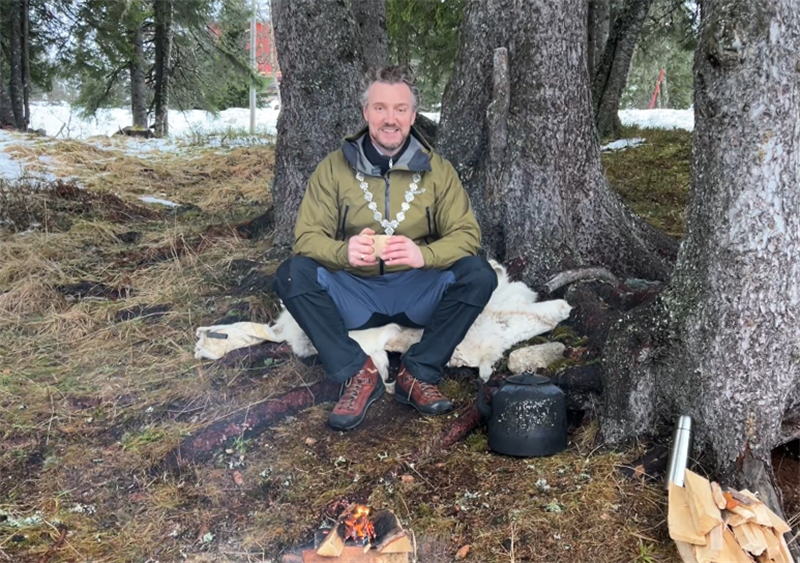 Fylkesordfører Tomas Iver Hallem sitter ved et bål. Han har på seg turklær og ordførerkjede og holder en kaffekopp i hånda.