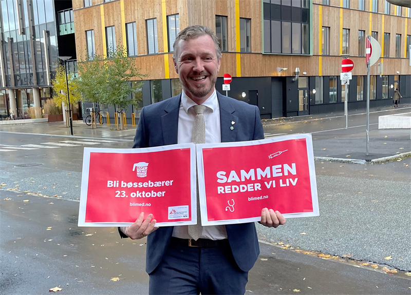 Fylkesvaraordfører Tomas Iver Hallem står utendørs og holder i to plakater med budskapene "Bli bøssebærer 23. oktober" og "Sammen redder vi liv"